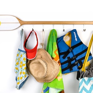 May include: A wooden paddle oar with hooks attached to the bottom, used as a wall-mounted rack. Hanging from the hooks are a swimsuit, a hat, a towel, a life vest, and a beach bag.