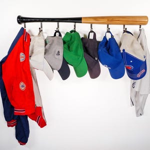 May include: A wooden baseball bat with hooks attached to it, used as a wall-mounted hat rack.  A red, white and blue baseball jacket, a white baseball cap, a green baseball cap, a grey baseball cap, a black baseball cap, a blue baseball cap, a white baseball cap with red and blue accents, and a white baseball jersey are hanging on the hooks.