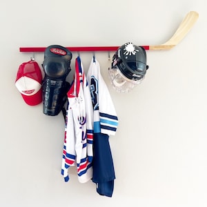 Puede incluir: Una camiseta de hockey roja, blanca y azul cuelga de un estante de madera roja con un casco de hockey, una espinillera de hockey y una gorra de béisbol.