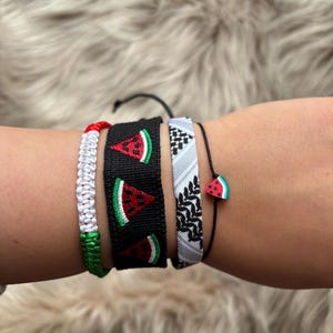 Op de afbeelding: Een verzameling handgemaakte armbanden. Eén armband heeft een watermeloenontwerp op een zwarte achtergrond. Andere armbanden zijn onder meer een wit en rood gevlochten ontwerp, een zwart-wit patroon en een kleine watermeloenbedel.