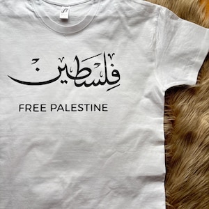 Könnte beinhalten: Weißes T-Shirt mit dem arabischen Wort für Palästina in schwarzer Schrift über dem englischen Text "FREE PALESTINE". Das Shirt hat einen Rundhalsausschnitt und kurze Ärmel.