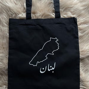 Könnte beinhalten: Schwarze Einkaufstasche mit einer weißen Umrandung des Landes Libanon und dem Wort "لبنان" in arabischer Schrift. Die Tasche hat schwarze Henkel und ein weißes "Samys Atelier" Logo unten rechts.