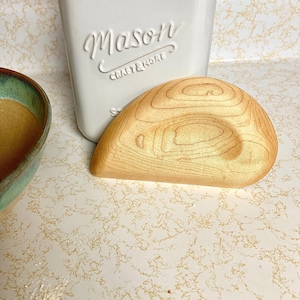 May include: A light-colored wooden dough scraper with a curved design. The scraper is next to a white container with the word "Mason" embossed on it. A ceramic bowl with a green and brown rim is in the background.