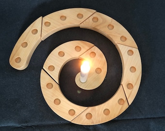 Può includere: Un calendario dell'Avvento in legno a forma di spirale con 24 sezioni individuali, ciascuna con un piccolo foro per una candela. Una singola candela bianca è accesa al centro della spirale.