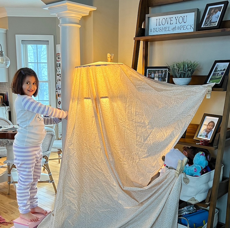 May include: A young girl wearing striped pajamas stands in front of a lampshade fort. The lampshade is covered in a white fabric with a floral pattern. The fort is built around a pillar in a room with a wooden floor. The sign above the fort reads "I love you a bushel and a peck."