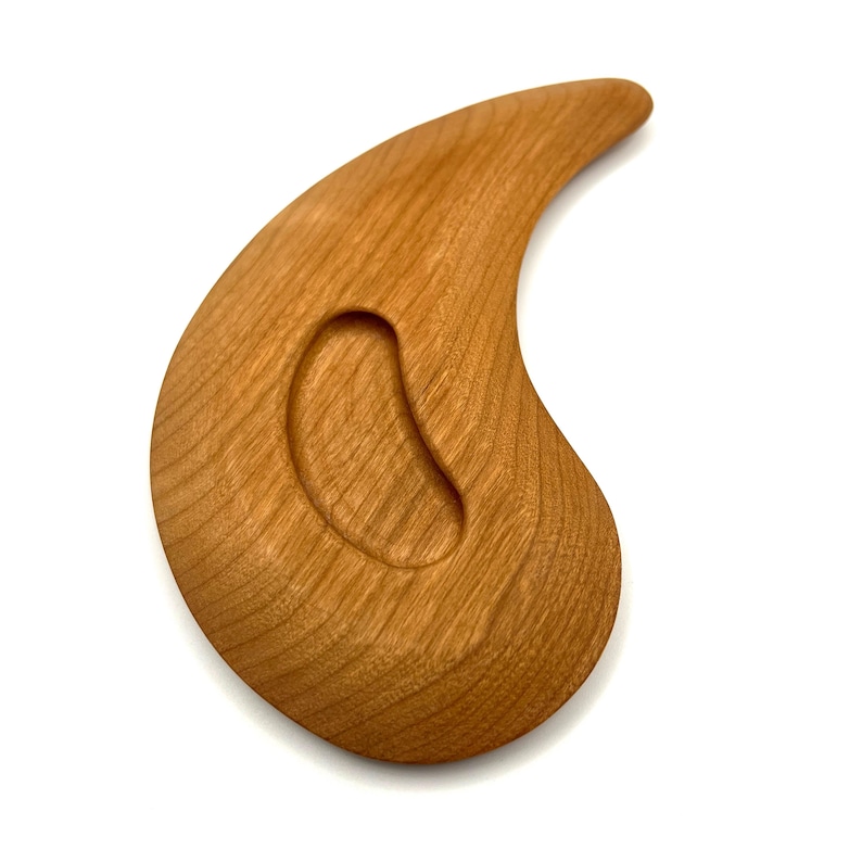 May include: A wooden massage tool, teardrop-shaped with a smooth, polished finish. It features a curved design with a rounded handle and an oval indentation. The wood grain is visible, displaying a warm, natural colour. The tool is likely used for therapeutic massage.