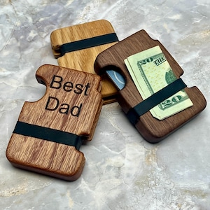 May include: Three wooden wallets with black elastic bands. One wallet is engraved with "Best Dad." Another holds a twenty-dollar bill. The wallets are made of different wood grains, including light and dark brown tones.