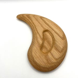 May include: A wooden massage tool with a curved, ergonomic design. The light brown tool features a smooth, polished finish and a carved indentation for grip. The wood grain is visible, adding to its natural aesthetic.