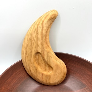 May include: A light-coloured wooden massage tool with a curved, ergonomic design. The tool has a smooth finish and a small, oval-shaped indentation. The wood grain is visible, and it rests on a dark brown, ceramic bowl.