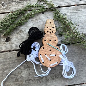 May include: Wooden snowman shaped lacing toy with white and black yarn and a needle. The snowman has a round body and a hat.