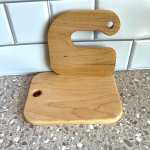 May include: Two wooden cutting boards, one shaped like a C with a hole in the top, and the other a rectangular shape with a hole in the center. Both boards are made of light wood.