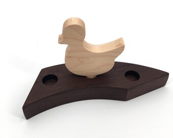 May include: A wooden duck figurine with a brown base. The duck is light brown and has a simple design. The base has two small holes.