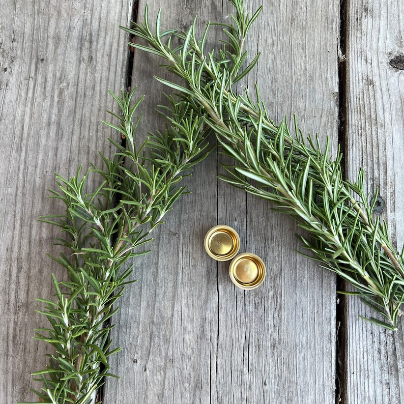 Pu&ograve; includere: Due tappi di bottiglia in metallo color oro su una superficie di legno con due rametti di rosmarino.