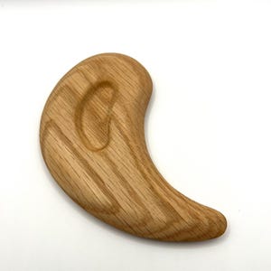 May include: A light brown wooden massage tool with a curved, ergonomic design. The tool has visible grain patterns and a smooth, polished surface. Its rounded shape is designed for bodywork and relaxation.