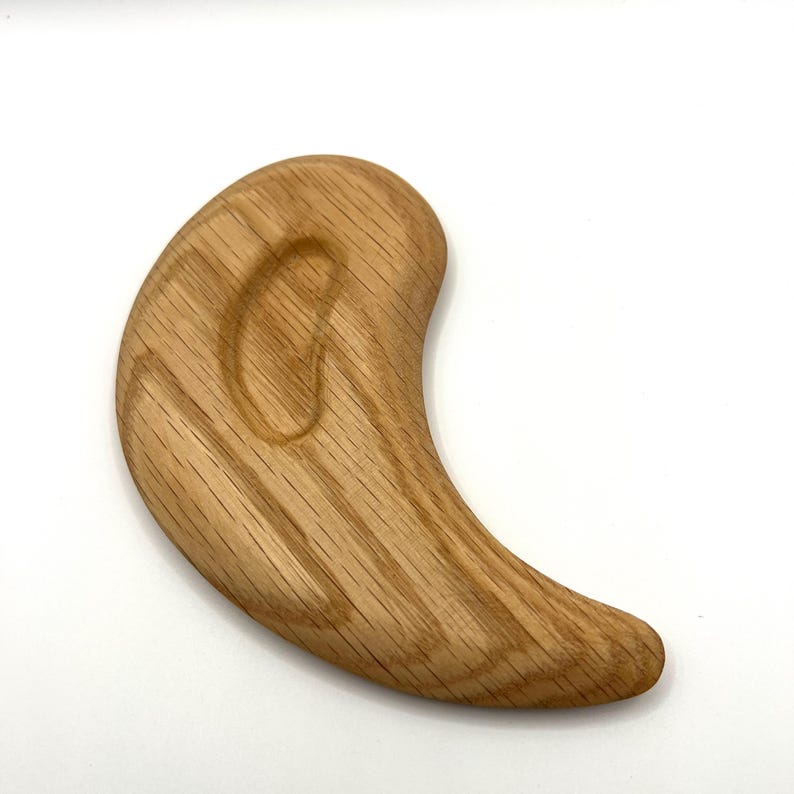 May include: A light brown wooden massage tool with a curved, ergonomic design. The tool has visible grain patterns and a smooth, polished surface. Its rounded shape is designed for bodywork and relaxation.