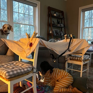 May include: A child in yellow and white striped pajamas plays in a fort made from white sheets and chairs. The fort is decorated with clothespins and a black strap. A dog is peeking out from behind a chair.