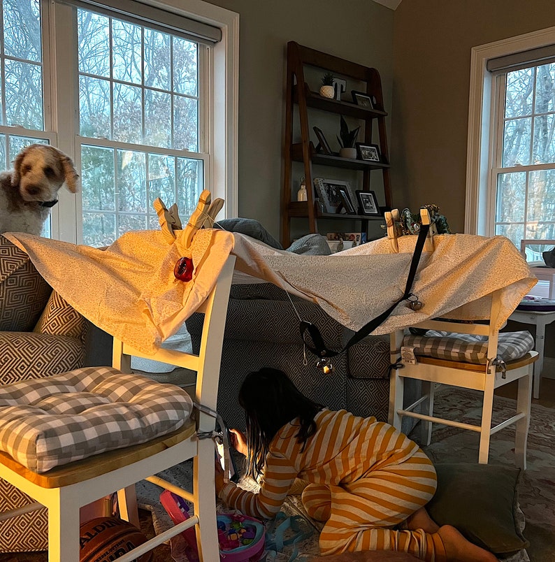 May include: A child in yellow and white striped pajamas plays in a fort made from white sheets and chairs. The fort is decorated with clothespins and a black strap. A dog is peeking out from behind a chair.