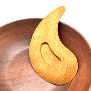 May include: A light-coloured wooden gua sha tool with a curved design and an oval cutout, resting on a brown bowl. The gua sha tool has a smooth, polished finish, and the bowl has a rustic, earthy appearance.
