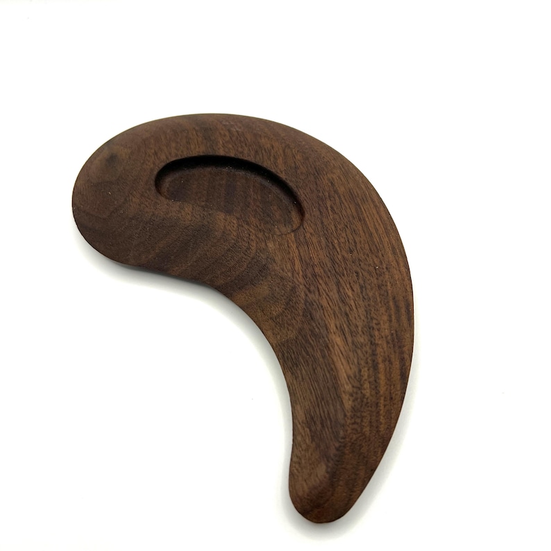 May include: A dark brown wooden massage tool with a curved, ergonomic design. It has a rounded handle with a recessed area and a smooth, polished finish. The wood grain is visible.