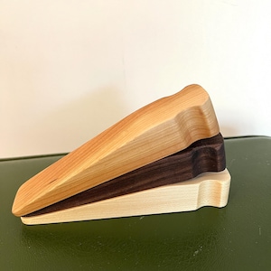 May include: Three wooden doorstops in different wood types: light wood, dark wood, and a lighter wood with a slightly darker hue. The doorstops are stacked on top of each other.