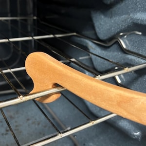 May include: A wooden oven rack puller with a curved handle and a cutout for gripping the rack. The puller is light brown and is inserted into the oven rack. The oven interior is dark blue.