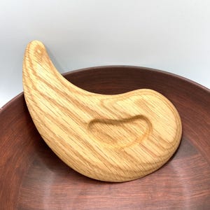 May include: A light-coloured wooden gua sha tool with a curved, ergonomic design. The tool has a smooth, polished finish and is resting on a dark brown ceramic bowl. The gua sha tool is designed for facial massage and skincare.