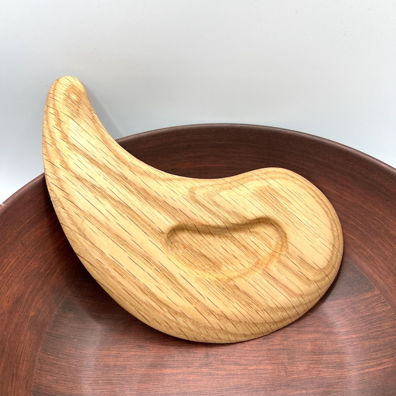 May include: A light-coloured wooden gua sha tool with a curved, ergonomic design. The tool has a smooth, polished finish and is resting on a dark brown ceramic bowl. The gua sha tool is designed for facial massage and skincare.