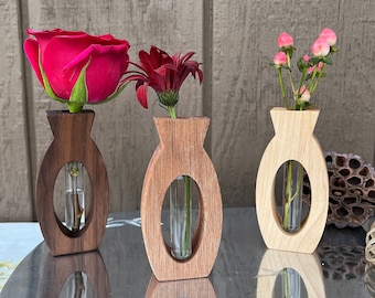 May include: Three wooden bud vases, each holding a single flower. The vases are oval-shaped with a clear glass tube in the center. One vase holds a red rose, another a dark red flower, and the third, pink berries.