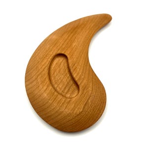 May include: A wooden massage tool, teardrop-shaped with a curved handle. The tool is made of light brown wood and has a smooth, polished finish. It features a small, oval-shaped indentation in the centre.