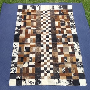 Puede incluir: Una alfombra rectangular de piel de vaca con un diseño de retazos. La alfombra presenta cuadrados de varios tonos de marrón, negro y blanco. Un patrón de tablero de ajedrez recorre el centro. La alfombra está sobre una superficie azul.