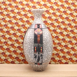 Puede incluir: Un jarrón de cerámica blanca con un esmalte craquelado y patrones geométricos en negro, rojo y naranja. El jarrón tiene forma ovalada y un cuello estrecho.