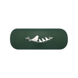Puede incluir: Un estuche oblongo de color verde oscuro con un adorno metálico plateado en el centro. El elemento decorativo muestra una fila de cuatro pájaros posados sobre una rama fina y curva, con el pájaro más grande situado a la izquierda.