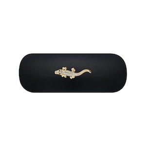 Puede incluir: Un estuche rígido negro mate de forma ovalada con un emblema de metal dorado en forma de caimán o lagarto centrado en la tapa. La figura del reptil está adornada con pequeños diamantes de imitación transparentes y brillantes.