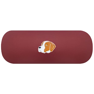 Puede incluir: Un estuche rígido para gafas de forma ovalada en color granate, con un gráfico central estilizado tipo esmalte de una cabeza de perro. La ilustración muestra pelaje marrón y blanco con una nariz negra y hocico blanco sobre el fondo rojo oscuro.