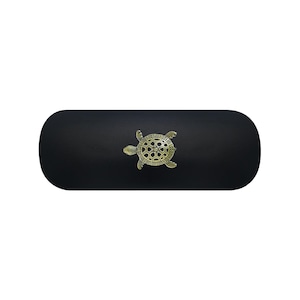 Pode incluir: Um estojo rígido oblongo de cor preta mate com um emblema de tartaruga em metal dourado centrado. O design da tartaruga apresenta padrões geométricos intrincados na carapaça e membros detalhados, criando um contraste com a superfície escura.
