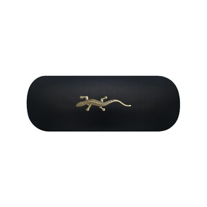 May include: A matte black, oblong-shaped hard case featuring a centered, metallic gold-toned lizard emblem. The case has rounded ends and a smooth, solid-colored surface, providing a clean and simple aesthetic for storage or protection.