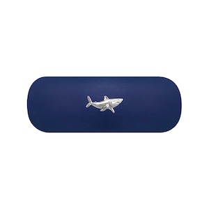 Könnte beinhalten: Ein dunkelblaues, längliches Hartschalenetui mit abgerundeten Enden. In der Mitte der Vorderseite befindet sich ein kleines, detailliertes, silberfarbenes Hai-Emblem aus Metall, das horizontal ausgerichtet ist und dessen Kopf nach rechts zeigt.