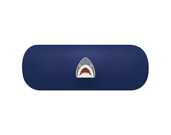 May include: A dark blue, oblong-shaped hard-shell glasses case featuring a centered graphic of a shark head. The shark illustration displays a grey upper body, a white underbelly, and an open mouth revealing sharp, dark red jagged teeth.