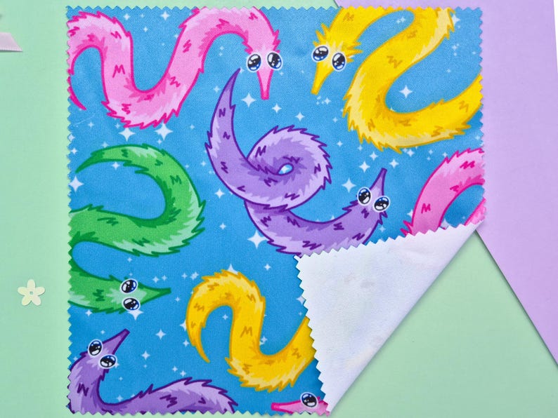 May include: A square fabric with a blue background featuring a pattern of colorful cartoon worms. The worms are in shades of pink, yellow, green, and purple, with white eyes and a starry background. The fabric is folded over, revealing a white underside.