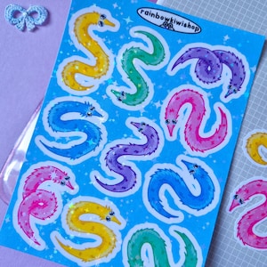 Könnte beinhalten: Ein Bogen mit bunten Wurmaufklebern in verschiedenen Farben, darunter Gelb, Grün, Blau, Lila und Rosa. Die Aufkleber haben einen glitzernden, holografischen Effekt und befinden sich auf einem hellblauen Hintergrund. Der Text "rainbowkiwishop" ist sichtbar.