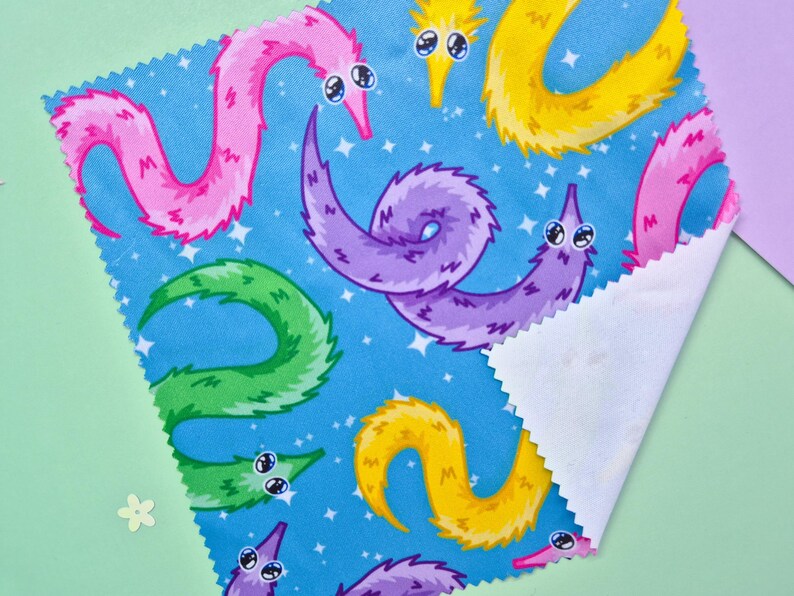 May include: A square piece of fabric with a blue background featuring a repeating pattern of colorful worm-like creatures. The worms are in shades of pink, purple, yellow, and green, with white eyes and a starry sky.
