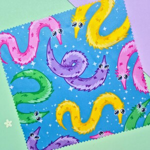 May include: A square fabric with a blue background featuring a pattern of colorful cartoon worms. The worms are in shades of pink, yellow, green, and purple, each with large, expressive eyes. The fabric is set against a backdrop of light green and purple.