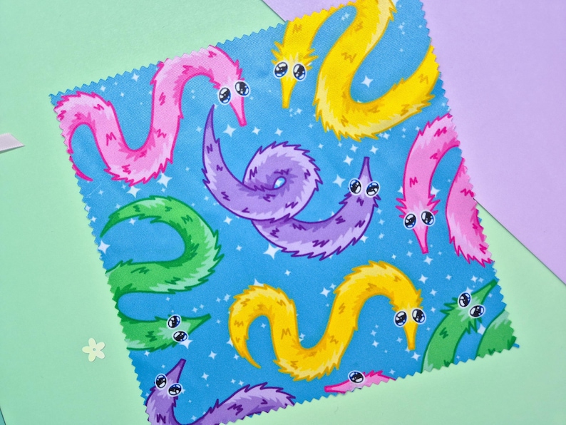 May include: A square fabric with a blue background featuring a pattern of colorful cartoon worms. The worms are in shades of pink, yellow, green, and purple, each with large, expressive eyes. The fabric is set against a backdrop of light green and purple.
