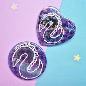 May include: Two holographic pins, one heart-shaped and one round, featuring a worm design. The pins have a purple and black checkered pattern with the text "It's not just a phase, mom."