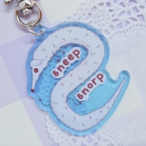Könnte beinhalten: Ein blauer Acryl-Schlüsselanhänger mit einem weißen, wurmartigen Wesen und den rosa Worten "sneep" und "snorp". Der Schlüsselanhänger hat einen silberfarbenen Verschluss.