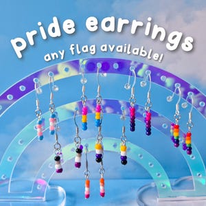 Könnte beinhalten: Pride-Ohrringe in verschiedenen Flaggen-Designs hängen an einem regenbogenförmigen Ständer. Die Ohrringe bestehen aus bunten Perlen. Der Text lautet "Pride earrings any flag available!"