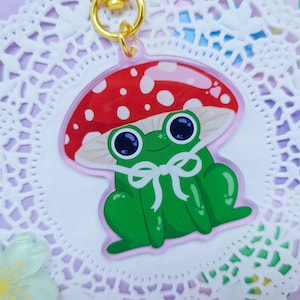 Könnte beinhalten: Ein grüner Frosch, der einen roten und weißen Pilz-Hut mit einer weißen Schleife trägt. Der Frosch sitzt auf einem weißen Spitzenhintergrund.