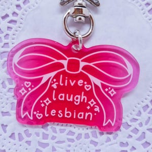 Può includere: Un portachiavi in acrilico rosa a forma di fiocco con la scritta bianca "live laugh lesbian". Il portachiavi ha un fermaglio color argento.