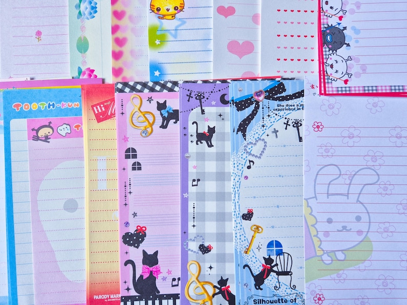 Puede incluir: Una colecci&oacute;n de art&iacute;culos de papeler&iacute;a coloridos, que incluyen blocs de notas y papeles decorativos. Los dise&ntilde;os presentan personajes de dibujos animados, notas musicales, gatos, corazones y patrones florales. La paleta de colores incluye rosa, azul, morado y amarillo.