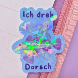 May include: A holographic sticker with a blue outline and the words "Ich dreh Dorsch" in blue text. The sticker features a fish design with purple, green, and yellow accents. The sticker is placed on a pink and purple background.